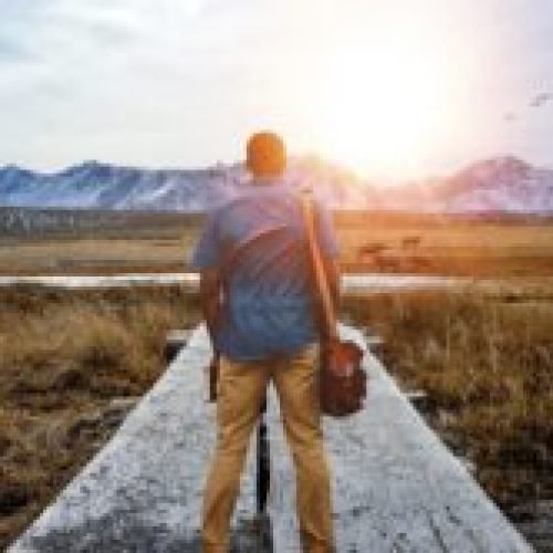 A shallow focus from behind of a male standing on a pathway in the middle of a grassy field with mountains in the distance