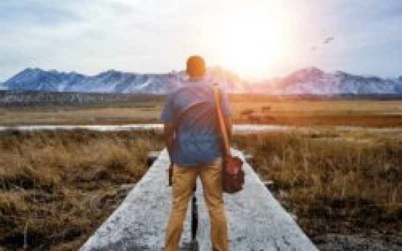 A shallow focus from behind of a male standing on a pathway in the middle of a grassy field with mountains in the distance
